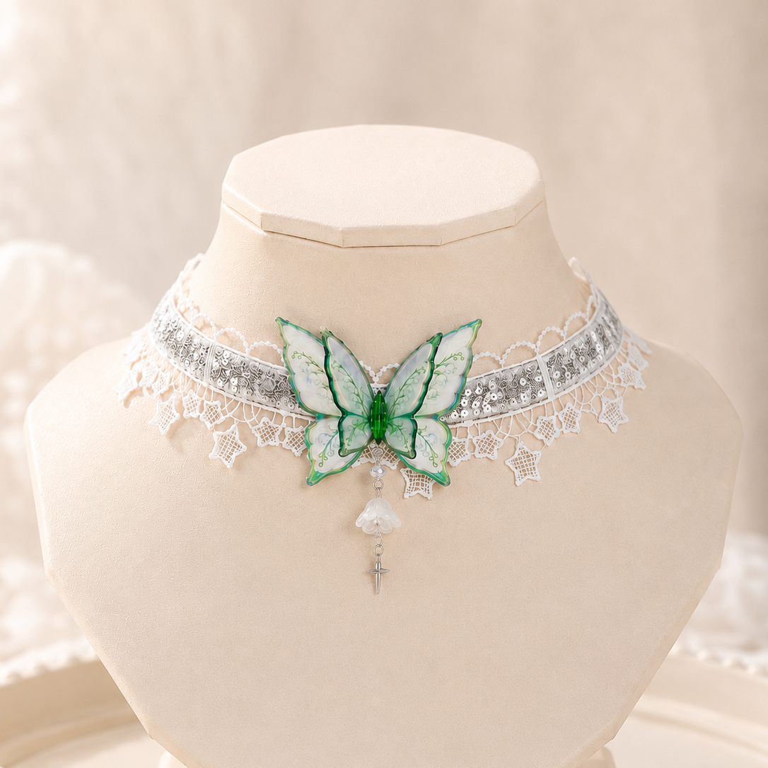 A delicate choker necklace displayed on a cream jewelry bust. The necklace features a silver-toned band with white lace trim, a central green and white butterfly motif, and a dangling white pearl-like bead with a silver cross charm.