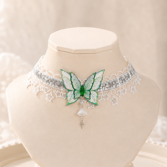 A delicate choker necklace displayed on a cream jewelry bust. The necklace features a silver-toned band with white lace trim, a central green and white butterfly motif, and a dangling white pearl-like bead with a silver cross charm.