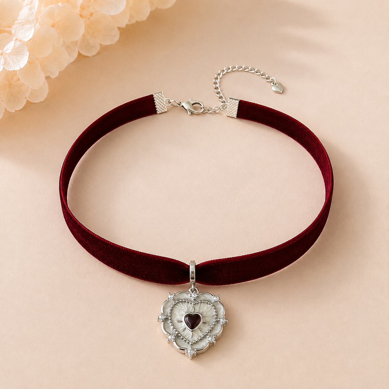 A velvet choker necklace in deep burgundy red with a silver-toned heart pendant featuring a small dark gemstone, displayed on a soft beige background with decorative flowers in the corner.