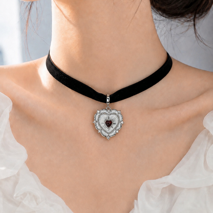A close-up of a model wearing a black velvet choker necklace with a silver heart-shaped pendant featuring a dark red gemstone.
