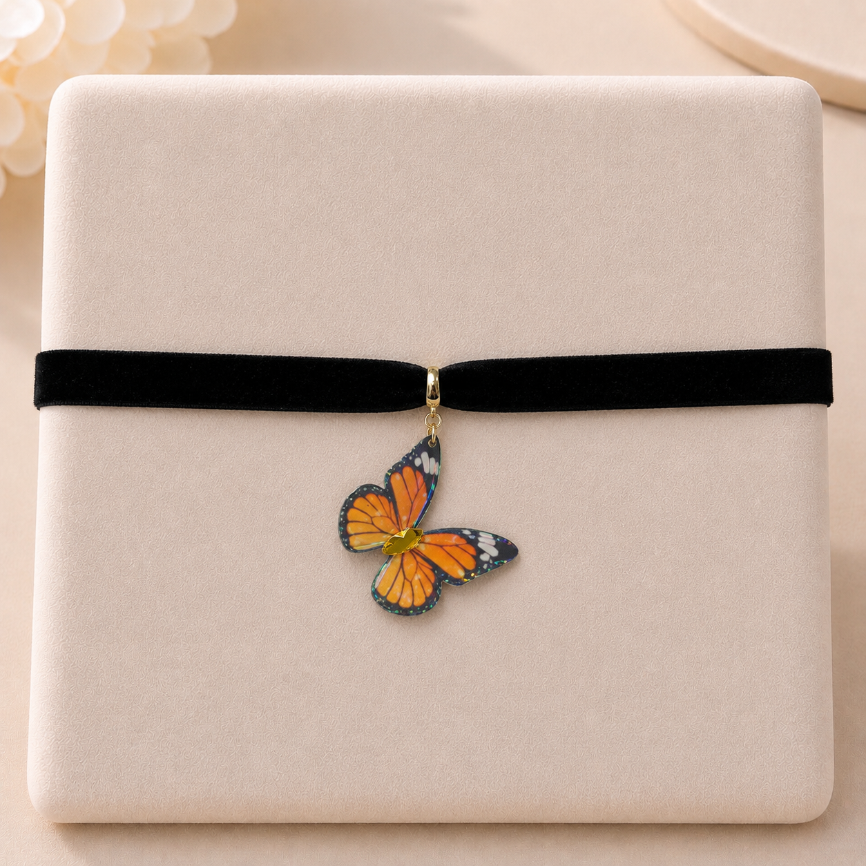 A close-up of a black velvet ribbon wrapped around a cream-colored textured box, with a small butterfly-shaped charm hanging from the center. The butterfly has orange, black, and white wings with a gold-toned metal clasp.