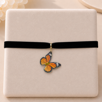 A close-up of a black velvet ribbon wrapped around a cream-colored textured box, with a small butterfly-shaped charm hanging from the center. The butterfly has orange, black, and white wings with a gold-toned metal clasp.