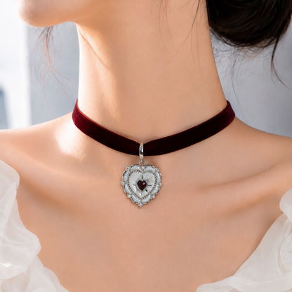 A close-up of a model wearing a dark red velvet choker necklace with a silver heart-shaped pendant featuring a dark red center stone.