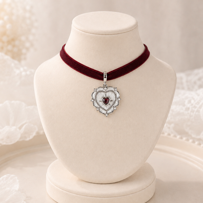 A heart-shaped silver pendant necklace with a red heart center, displayed on a cream-colored jewelry bust. The necklace is paired with a deep burgundy velvet ribbon choker, set against a soft, warm, white lace background.