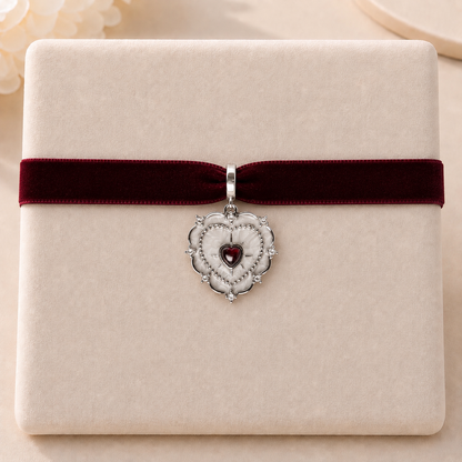 A silver heart-shaped pendant charm with a small red heart in the center, displayed on a cream-colored gift box tied with a dark red velvet ribbon.
